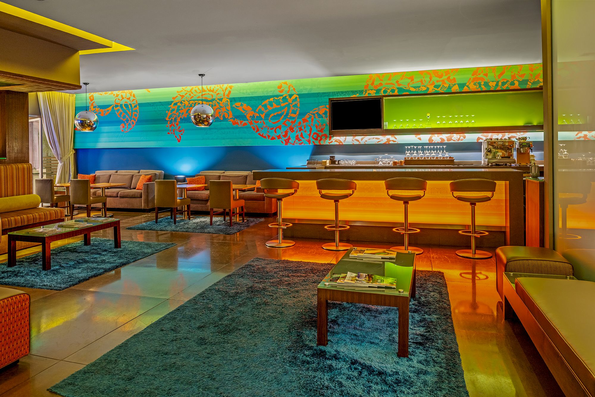 A modern lounge with a bar, colorful lighting, bar stools, couches, a television, and vibrant wall art.