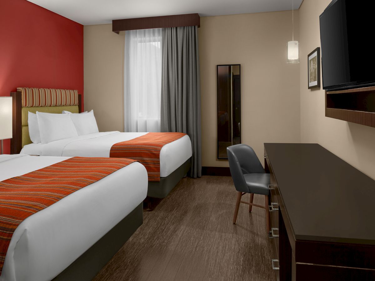 The image shows a hotel room with two beds, a desk with a chair, a wall-mounted TV, a mirror, and a window with curtains.