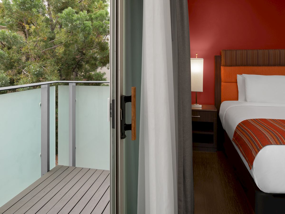 A modern hotel room with a double bed and red accents is adjacent to a small balcony with glass railing and a view of trees.