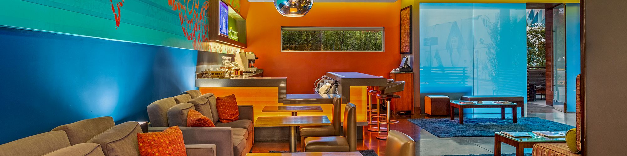 A colorful lounge with sofas, wooden tables, bright lighting, and decor. Walls blend blue, orange, and green hues, creating a vibrant atmosphere.