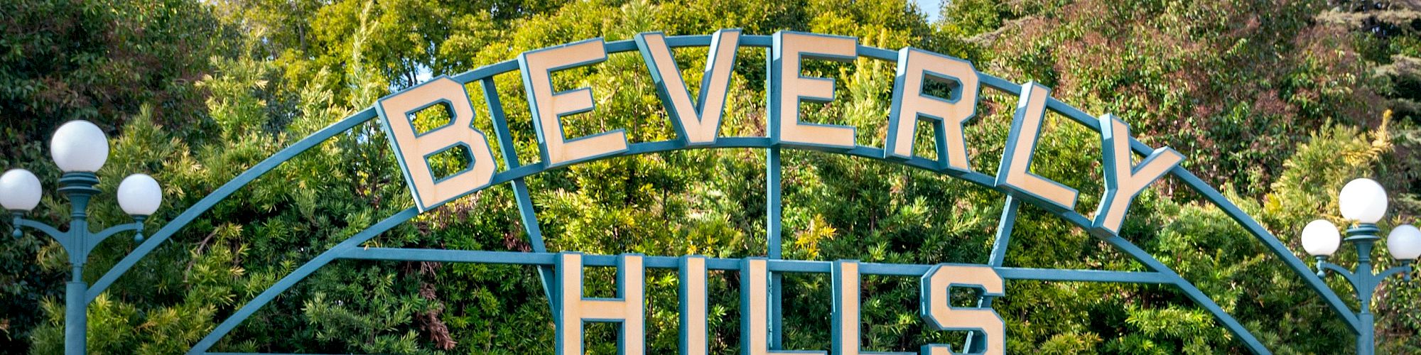 A sign reading “Beverly Hills” is surrounded by lush greenery and decorative lights, near a small body of water.