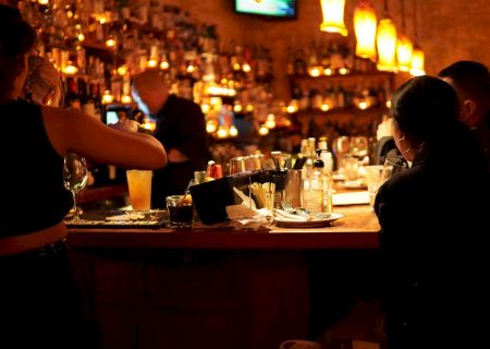 A warmly lit bar with patrons seated, bottles and glasses on the counter, and a bartender pouring drinks amidst a cozy atmosphere.