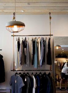 The image shows a clothing store interior with racks of clothes, hanging lights, and a person browsing in front of a mirror.