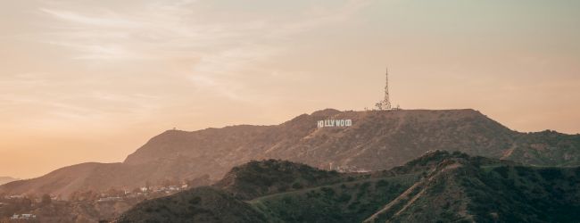 The image shows a landscape view of the Hollywood Hills with the famous Hollywood Sign in the distance at sunset.