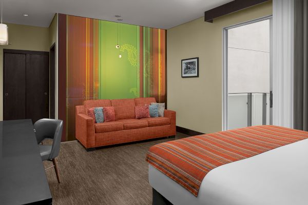 A modern hotel room features a bed with an orange-striped cover, an orange sofa, and a desk with a chair, with a balcony access.