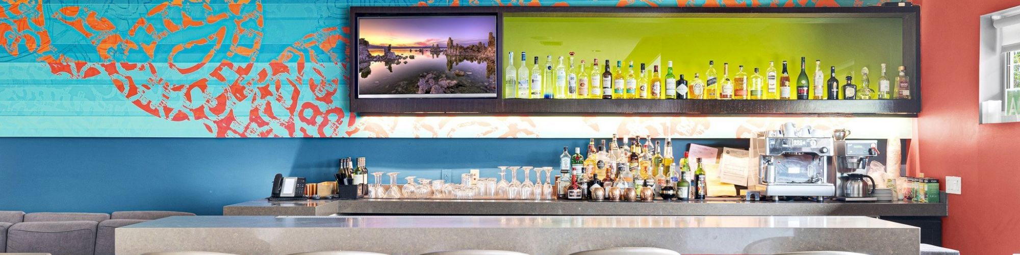 A modern bar with colorful decor, five stools, a TV, and various bottles on display against a vibrant background wall.