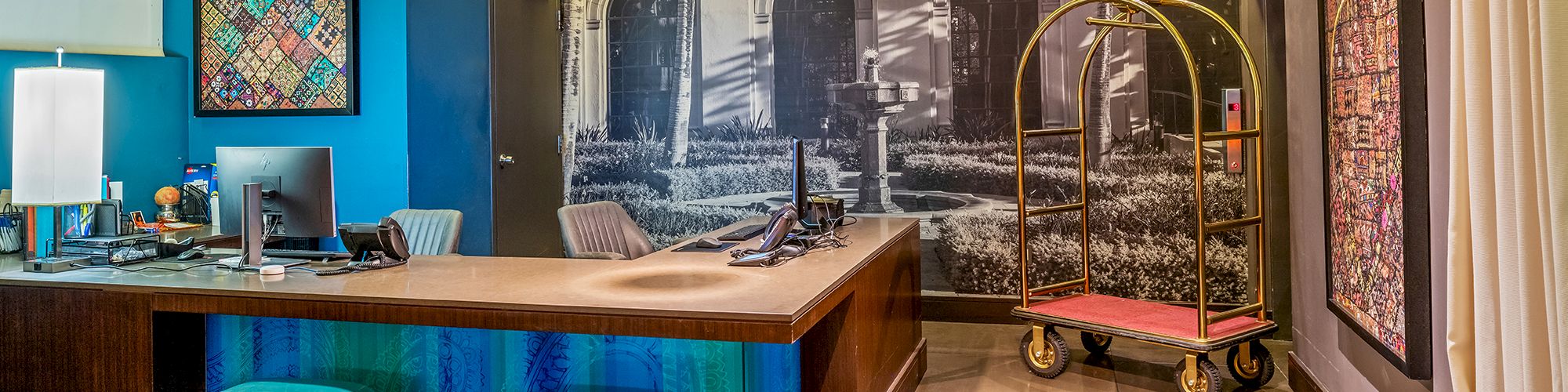 A modern hotel reception area features a desk, seating, wall art, and a luggage cart under ambient lighting.