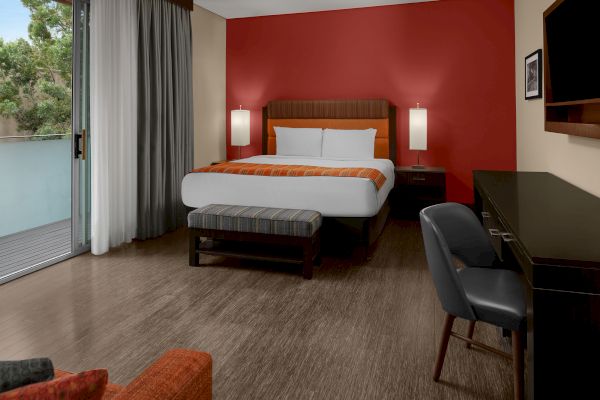 A modern hotel room with a bed, desk, chair, and a balcony. The room has red and cream walls, wooden flooring, and a large window.