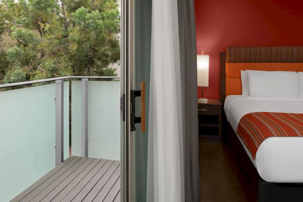 The image shows an open balcony door leading to a wooden deck, with a modern bedroom featuring a bed and orange accents visible inside.