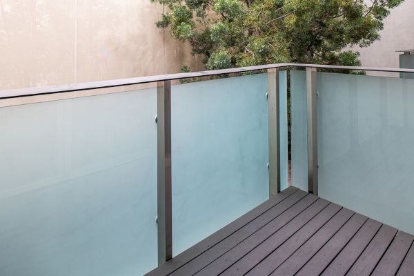 A small balcony with frosted glass panels and metal railings, featuring wooden flooring and a tree visible in the background.