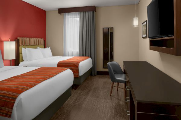 The image shows a hotel room with two beds, a TV, a desk, a chair, and a mirror under soft lighting.