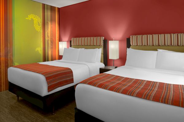 The image shows a hotel room with two neatly made double beds, red-striped bed runners, modern lamps, and colorful wall decor.