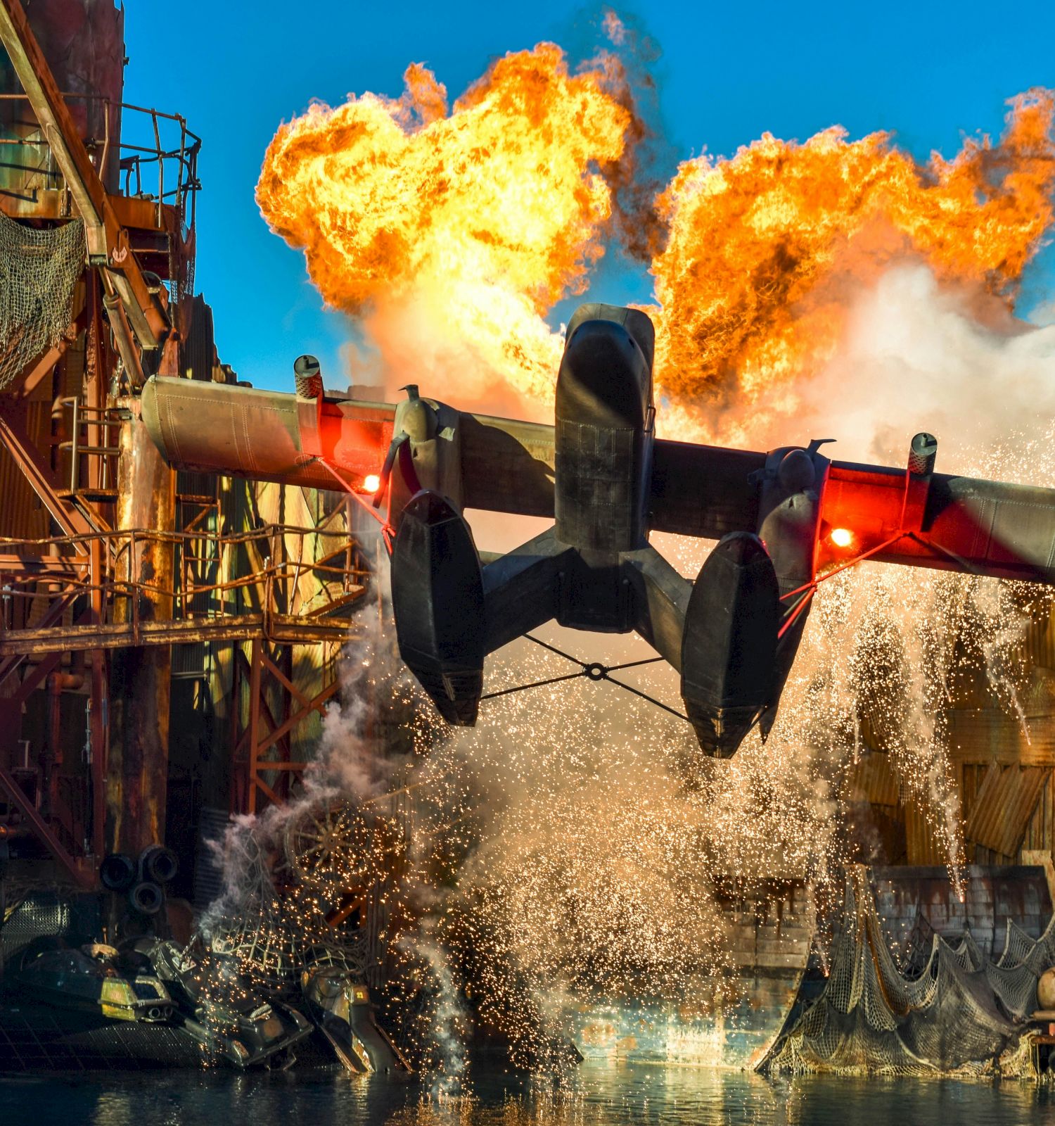 A seaplane dramatically lifts off from water amid fiery explosions and industrial structures in the background.