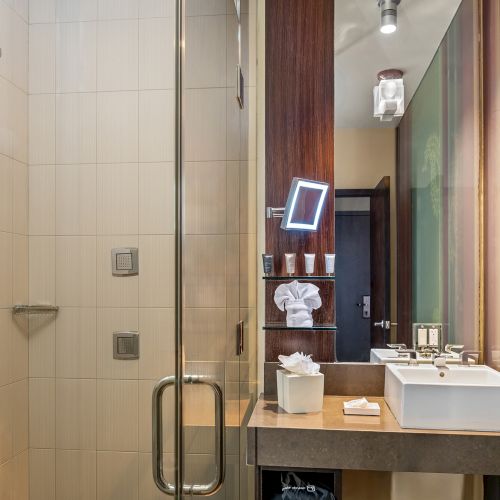 A modern bathroom with a glass shower, square sink, mirror, and toiletries on the counter. Walls feature a mix of tiles and wood accents.