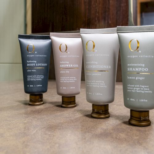 Four small bottles of bath products labeled body lotion, shower gel, conditioner, and shampoo on a bathroom counter.