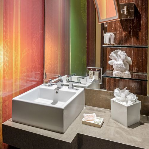 A modern bathroom with a square sink, colorful walls, mirror, towels, soap, and a tissue box on the counter.