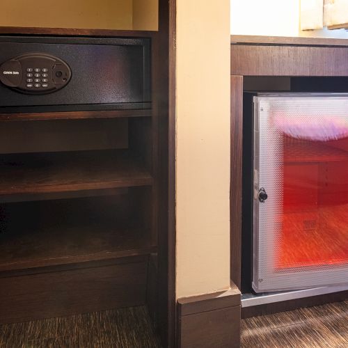 The image shows a hotel room setup with a safe on a shelf and a mini fridge with a transparent door displaying drinks inside.