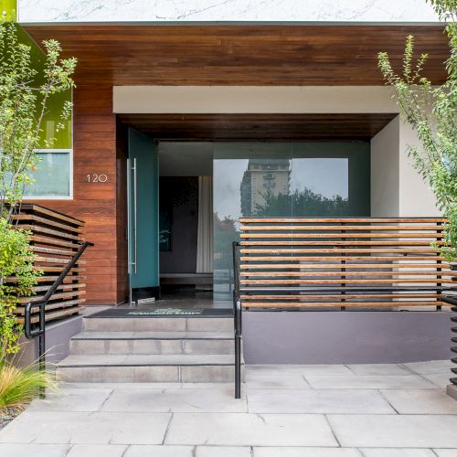 The image shows a modern house entrance with wooden accents, a blue door, plants, and a small staircase leading up to the entryway.