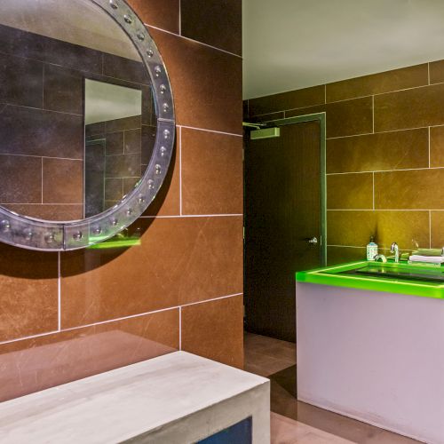 The image shows a bathroom with a round mirror and a sink with a glowing green edge, set against brown tiled walls and a closed door.