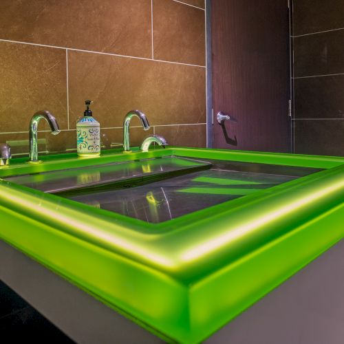 A modern bathroom sink with green LED lighting, two faucets, and a soap dispenser on a counter against brown tiled walls.