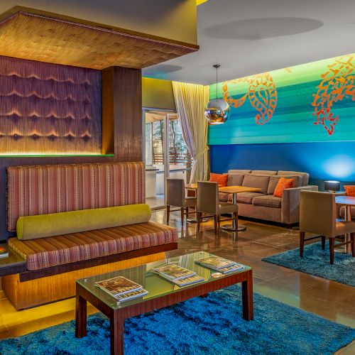 A modern lounge with colorful sofas, tables, and artistic wall designs. Pendant lights hang from the ceiling to brighten the space.