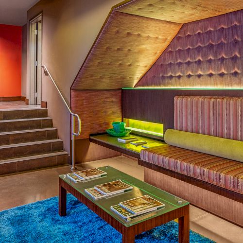 Modern seating area with striped cushions, a green table, magazines, and a blue rug. Stairs and elevator are in the background.