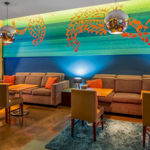 A modern lounge with sofas, tables, orange cushions, decorative wall art, and pendant lights, featuring a vibrant, colorful design.