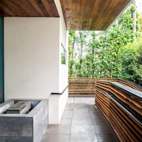 Modern outdoor patio with a minimalist design, featuring wooden accents, tiled flooring, and lush greenery in the background.