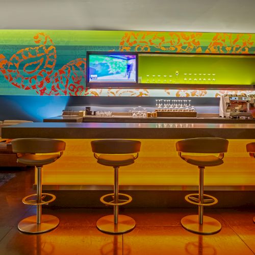 A modern bar with five stools, colorful wall art, a TV, and a coffee machine, creating a vibrant and cozy atmosphere.