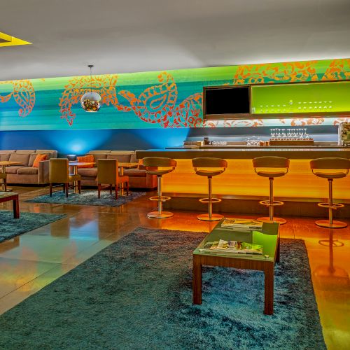 A modern bar with colorful decor, stools, sofas, and blue rugs. There’s a flat-screen TV and abstract art on the wall, creating a cozy vibe.