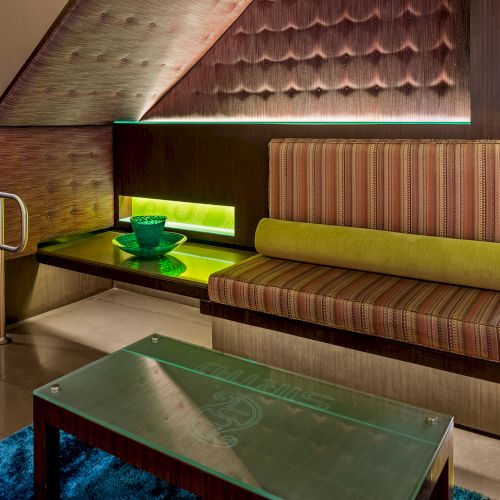 The image shows a modern seating area with striped cushions, a green-glass table, and a staircase, with subdued lighting and decor.