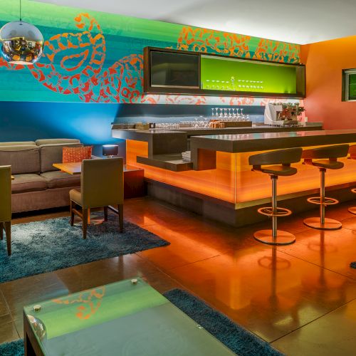 A modern bar with vivid colors, featuring a sofa, tables, bar counter with stools, and bright wall art in a cozy setting.