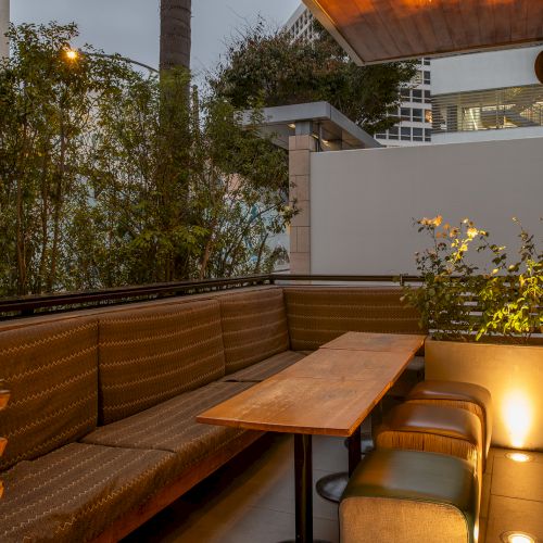 An outdoor patio with a wooden table, cushioned seating, plants, and city buildings in the background, lit by ambient lighting.