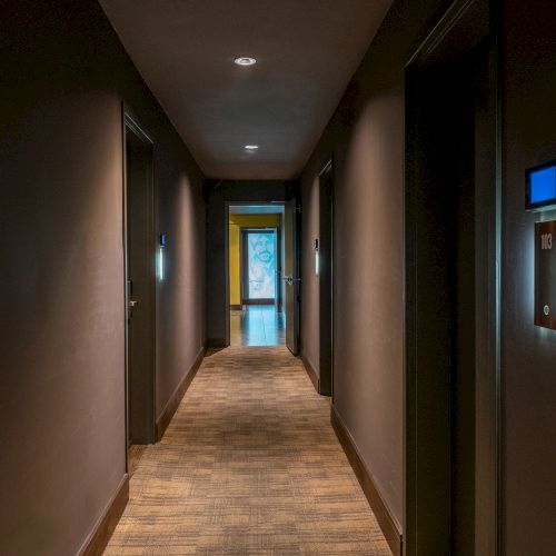 A dimly lit hotel hallway with closed doors on either side and a softly illuminated exit at the end of the corridor.