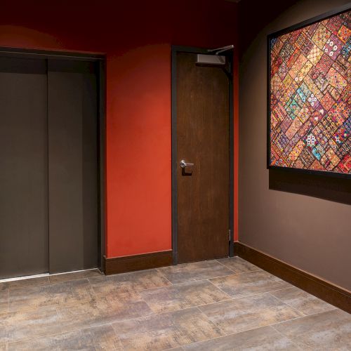 The image shows an elevator door, a wooden door, and a colorful framed artwork on a wall with a red background.