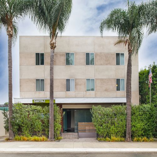 A modern building with a beige facade, surrounded by palm trees and greenery under a clear blue sky.