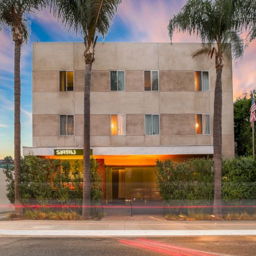 A modern building with large windows and palm trees in front, under a colorful sky at sunset. The scene captures a serene urban moment.