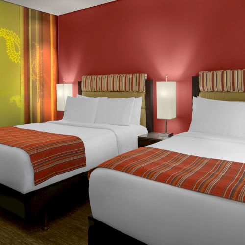 The image shows a hotel room with two neatly made beds, red walls, striped headboards, and modern bedside lamps.