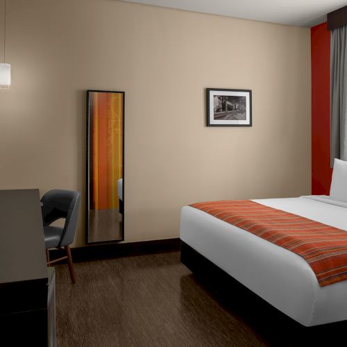 A modern hotel room with a large bed, wall art, a full-length mirror, desk, chair, and a TV. Light streams in through a window with curtains.