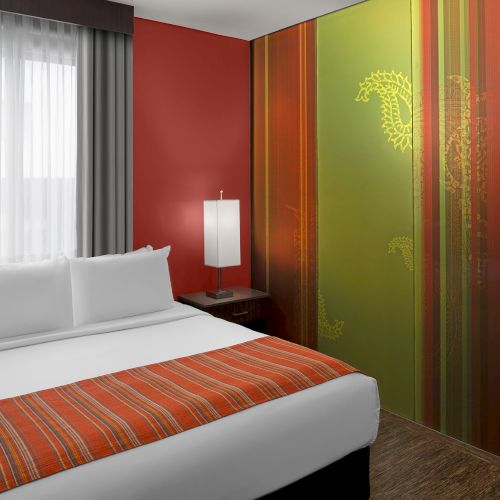 The image shows a modern hotel room with a double bed, vibrant wall art, and a bedside lamp in a cozy setting.