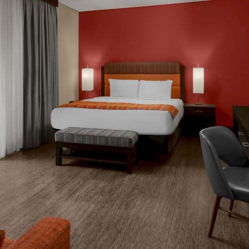 A modern hotel room with a bed, desk, chair, and red accent wall, adjacent to a sliding door leading to a balcony with trees visible outside.