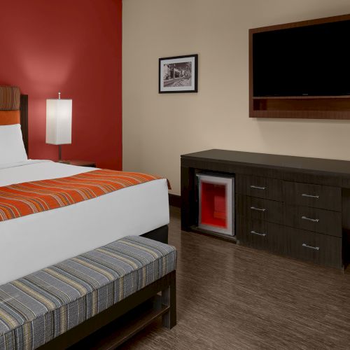 The image shows a hotel room with a bed, TV, desk, chair, mini-fridge, wall art, and a bench, featuring a red accent wall.