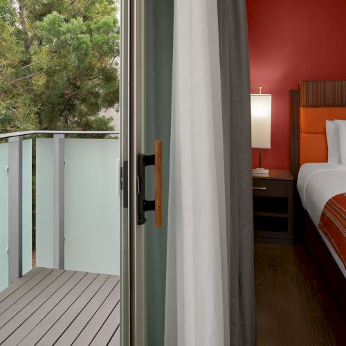 The image shows a hotel room interior with a bed next to a sliding door leading to a small balcony overlooking some trees.