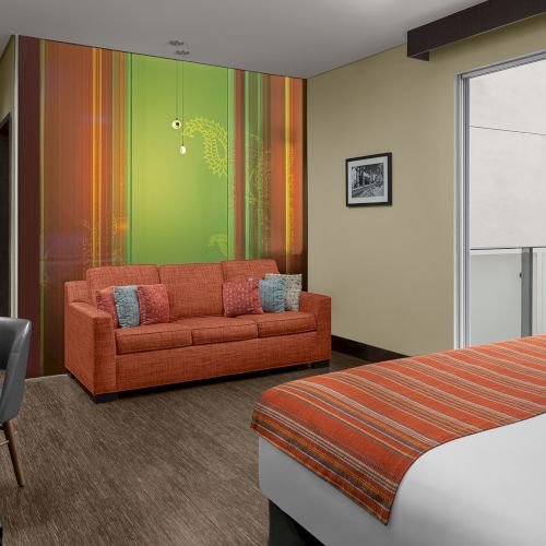 A modern hotel room with a bed, an orange sofa, a desk, and a colorful wall design, complemented by a small balcony area.