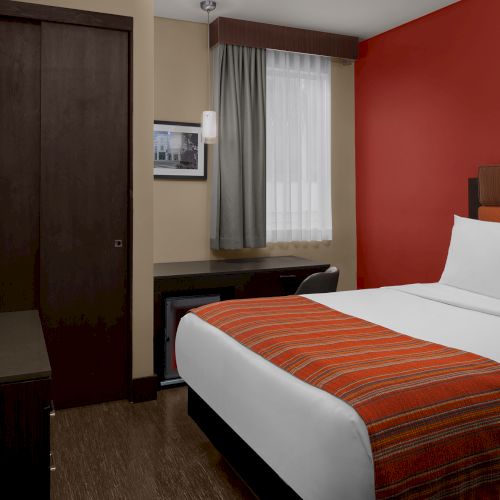 A cozy hotel room with a bed, red accent wall, TV, wardrobe, desk, and window with curtains. Warm lighting creates a welcoming ambiance.