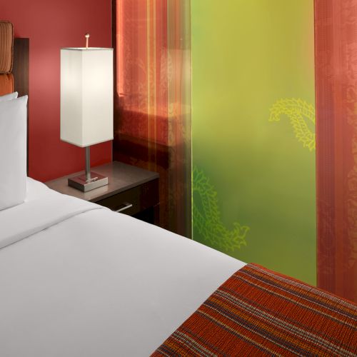 A neatly made bed with white linens, a nightstand with a lamp, and colorful curtains with green and red tones are in the room.