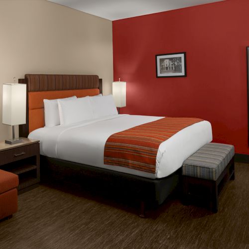 The image shows a hotel room with a large bed, red accent wall, armchair, desk, and mirror, creating a cozy atmosphere.