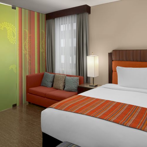 A hotel room with a bed, striped orange accents, a sofa, a modern lamp, and decorative glass panel, next to a window with curtains.