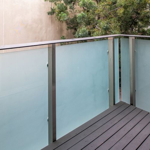 A balcony with frosted glass panels and a metal frame, surrounded by trees, with a wall in the background.