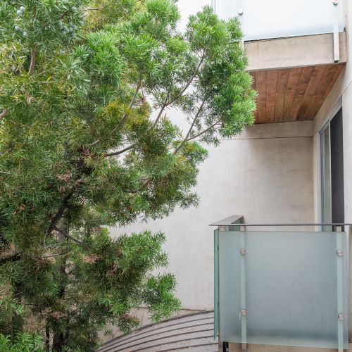 A small balcony with a frosted glass railing is adjacent to a building, next to a lush green tree under a cloudy sky.